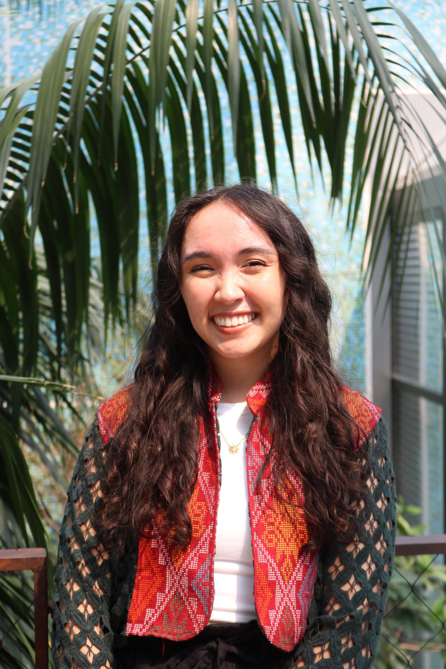 April, a light tan Southeast Asian person with loose, brown curls. April is wearing a jacket made from red and green traditional Philippine fabric and a white shirt. Behind April is a large, tropical tree and a blue wall.