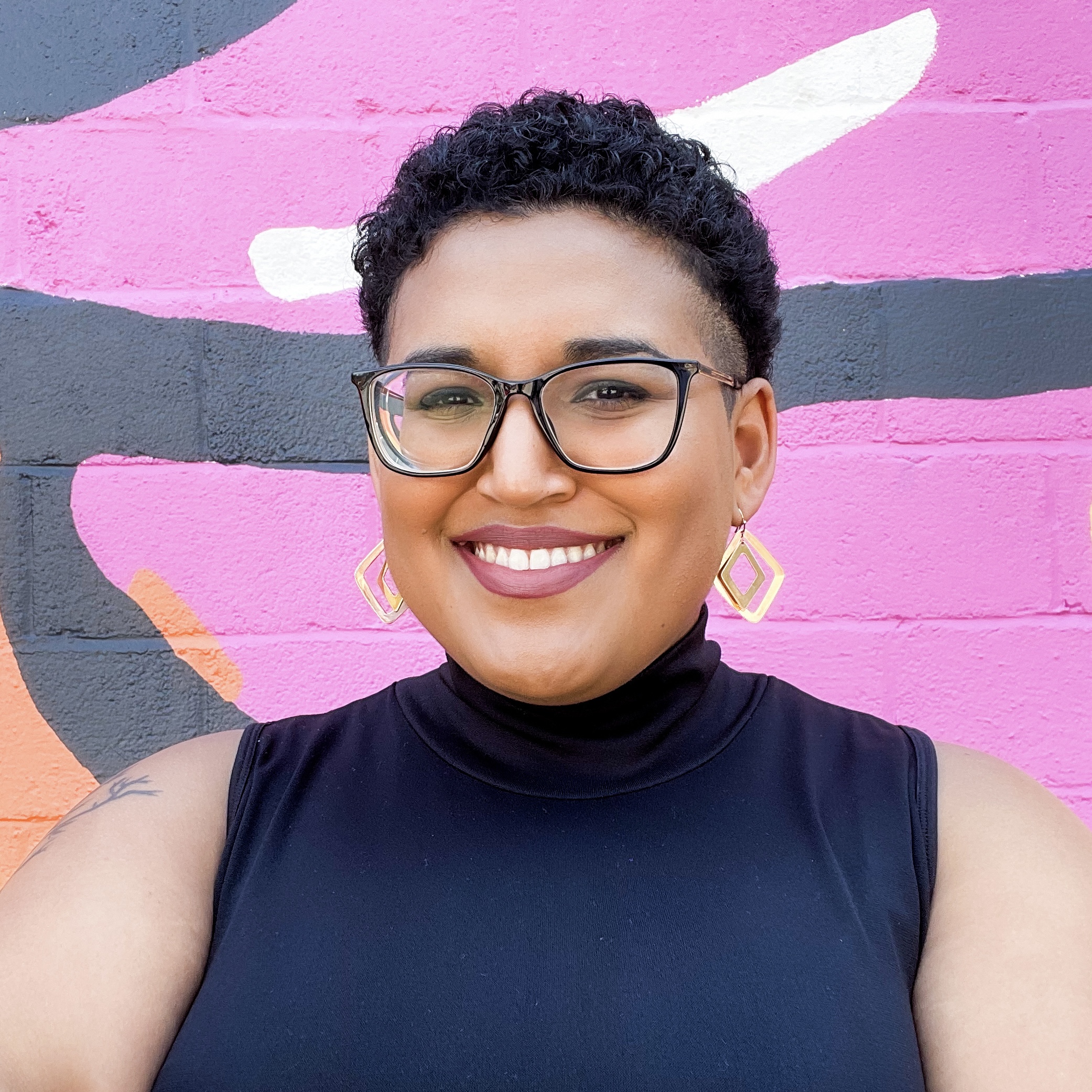 A Black person of Latinx descent with short curly hair smiles in front of a vibrant mural with pink, orange, and black abstract shapes. They are wearing glasses, geometric earrings, and a black sleeveless top.