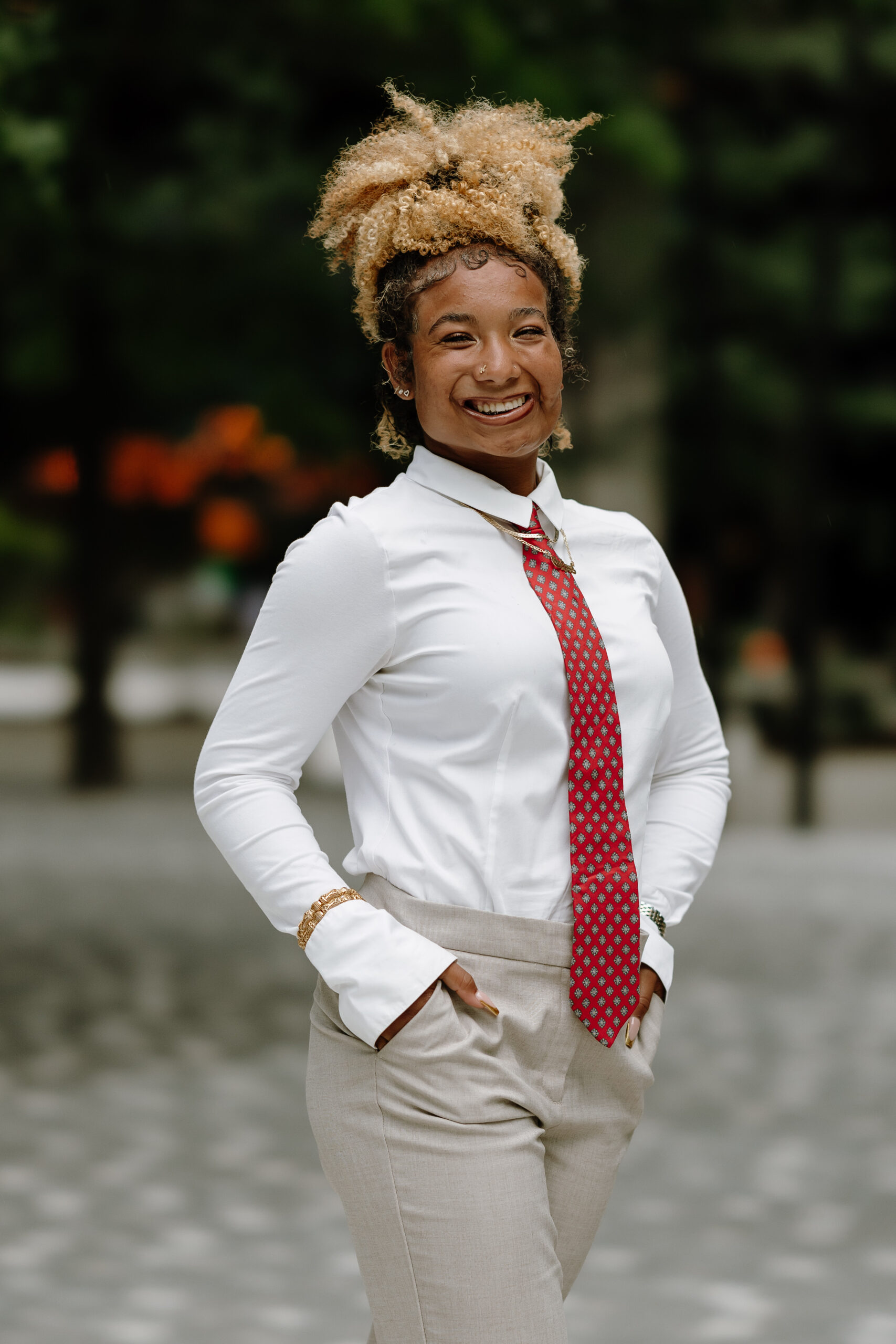 A Black female with blonde curly hair tied up smiling at the camera with her hands in her pocket. She is wearing a white long sleeved button down shirt with a red tie with white stars, gold bangles, and gray-tan pants.