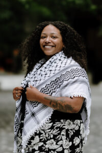 Headshot of Alyssa Christopher, a Black biracial person with long curly brown hair and tattoos on their forearm. They are smiling at the camera and wearing a black top, a black and white floral skirt, and a black and white keffiyeh.