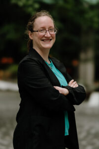 Headshot of Ann Johnston. A white person with dark-blond hair tied back and glasses. She is smiling, and wearing a teal top with a black blazer.