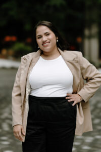 Light-skin Latina woman with shoulder-length brown hair and brown eyes, standing outside with left hand on waist wearing a white top, black pants, and light brown blazer.