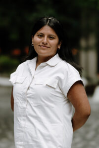 The image shows a woman with long dark hair, tan skin, and warm brown eyes smiling at the camera. She wears a white short-sleeved button-up shirt-dress with two front pockets. She stands in a park with blurred greenery as her background.