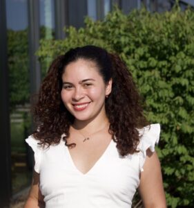 Emily Gonzalez is a Latinx woman with curly dark brown hair smiling at the camera. She is wearing red lipstick and a white short-sleeve blouse.