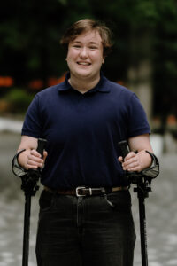 Headshot of Emmett Lockwood, a white passing white and indigenous man with brown hair wearing a blue polo, a brown belt with embroidered designs and black jeans. They are smiling and using two forearm crutches.