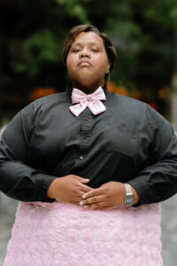 African-American male, with ear length curled brown hair wearing Ankh earring on left ear. Wearing black long-sleeved button down shirt, with “Hello Kitty” bowtie. Arms positioned at waste with hands placed on top hem of pink roses skirt.