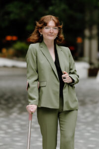 Katie, a white femme with a blunt auburn bob and gravity-defying bangs smiles slightly for the camera. She is bespectacled in vaguely hexagonal frames, donning an olive green suit and a can-do attitude.