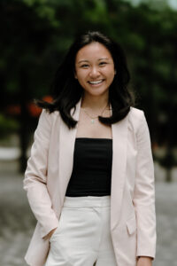 A Taiwanese and Vietnamese American woman with shoulder-length and curled ends, a jade necklace, silver studded earrings, a blush pink blazer with a black ribbed top, off-white colored trousers with her right hand in her pocket posing in front of a blurred background of trees