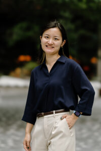 An Asian woman who is smiling with Black hair and yellow highlights. She is wearing a navy blue work blouse and beige trousers with a brown belt.