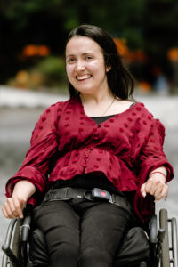 Maria is a light complected latina with straight brown hair. She is wearing a burgundy polka dot shirt with black jeans and is sitting in a manual wheelchair with a seat belt.