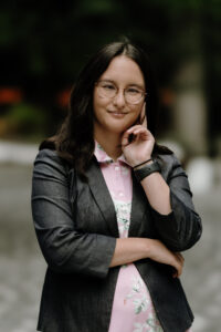 A mixed race girl with Vietnamese features, light tan skin, and long brown hair that goes past her shoulders wears dark red cat eye glasses smiles at the camera. She looks determined. She is wearing a burgundy red dress that is a mock turtleneck and a black blazer. The background is blurred greens and tans like she is standing in a forest.