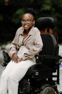 a picture of Nila Morton who’s a Black woman. She’s sitting in her black power wheelchair. She has dark brown twists which are in a high bun. She’s wearing black frame glasses with a beige long sleeve button down top. She’s wearing beige slacks. She has on small gold hoop earrings.