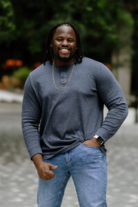 Ozzie (Black Male with locs) dressed in a navy blue shirt, gold chain and Jeans.