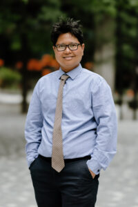 An Asian male wearing black glasses, light blue long-sleeve shirt, light maroon tie, and navy pants smiles and poses with his hands in his pant pockets