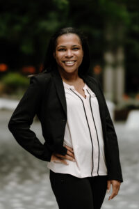 Headshot of Vanessa Sanders (she/they), a Black and Indigenous woman with brown skin and straight dark brown hair. She is wearing a pale pink blouse, a black blazer, and black pants.