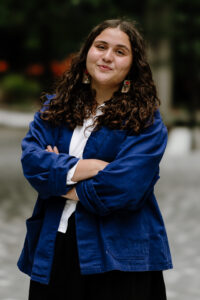 Zofia, a light-skinned, female presenting person is pictured smiling at the camera with their arms crossed. They have medium length, dark brown curly hair and brown eyes. They are wearing rectangular abalone earrings, a white button down, dark blue coat, and a black skirt. The background of the photo, a grey pavilion with green foliage and orange flowers, is blurred.