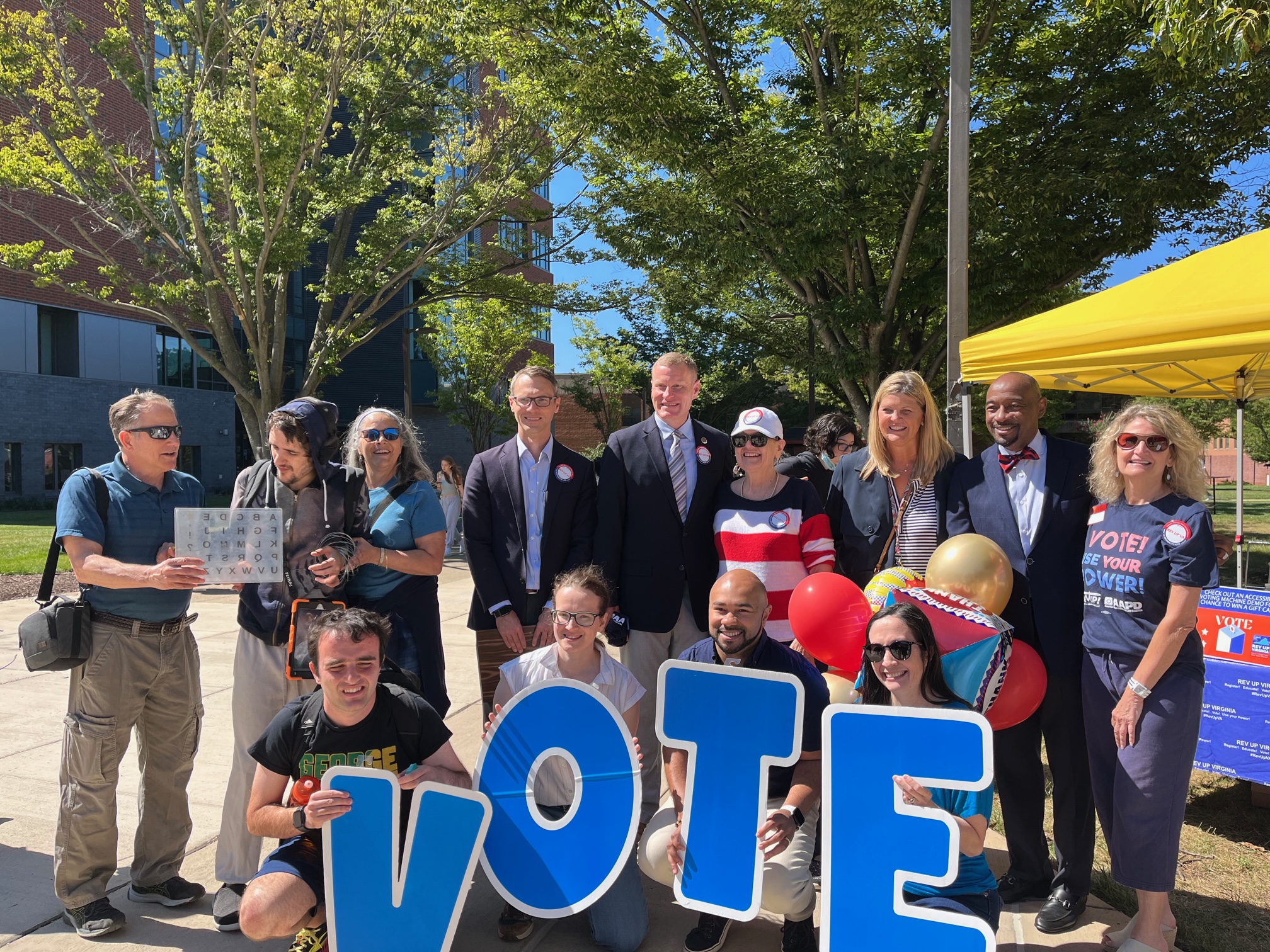 A group of people outdoors posing with large blue letters spelling "VOTE." The group includes men and women of different ages, with some holding balloons and signs. They are standing in front of a yellow tent, with trees and buildings in the background.