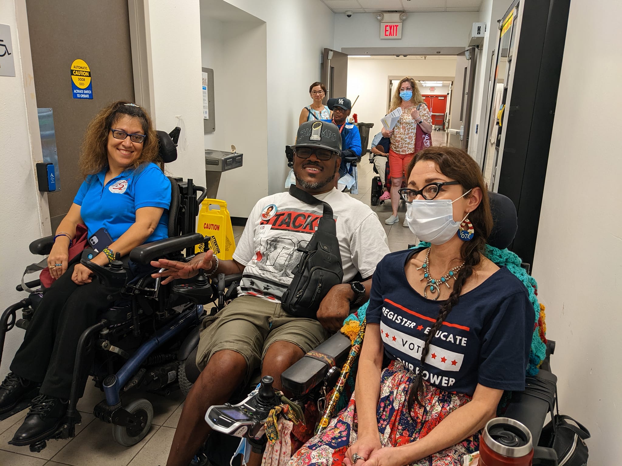 A group of three individuals in wheelchairs in a hallway. The person on the left is a woman with brown hair wearing glasses and a blue polo shirt. The person in the middle is a man wearing a white t-shirt, sunglasses, and a cap. The person on the right is a woman wearing a mask, glasses, and a dark blue shirt that reads "Register Educate Vote Power."