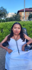 Fabiola, a brown woman with long black hair, is outside on a bright day. She wears a white dress, a bolo tie, and smiles at the camera. She is sitting on a black power wheelchair and wearing a red bracelet.