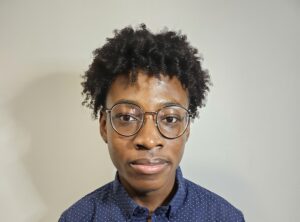 Jaydon is Afro-Trinidadian with black curly hair and wears glasses. He has on a blue t-shirt with white polka dots.