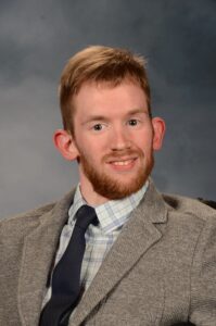 A white male with dirty blonde hair and a beard wearing a grey suit with a navy blue tie and a white and blue plaid button-down shirt.