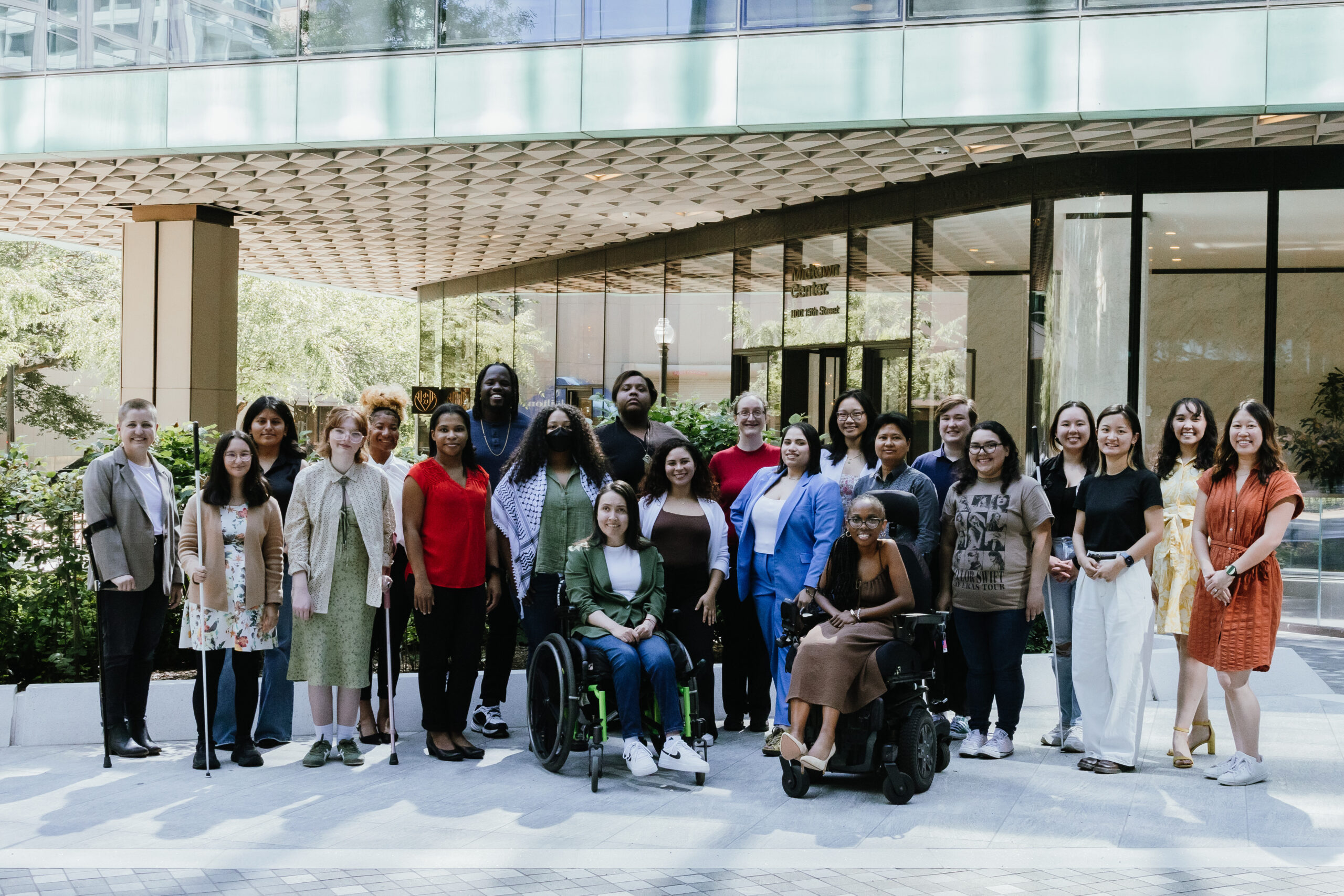 This image shows a diverse group of people standing together outside a modern building with large glass windows. The group includes men and women of various ages, ethnicities, and abilities, with two individuals in wheelchairs. The setting appears to be a professional or formal gathering, with the group posing for a group photo. The background features greenery and a paved area, contributing to the serene, outdoor environment.