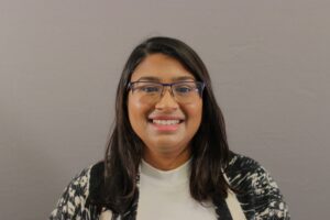 Shruti, a brown women with black hair just past her shoulders, is in front of a gray backdrop. She wears square glasses with a navy blue rim over the top and sides. She is smiling at the camera. She also is wearing a black and white marbled shawl over a white turtleneck shirt.