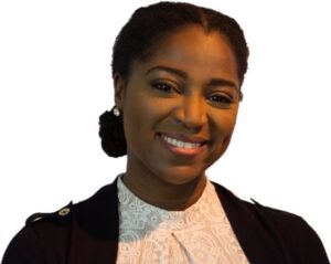 Image of a brown skinned woman with black hair braided in two braids going to the back into a tight bun, wearing a white laced blouse, and navy blue sweater. She is smiling with her head slightly tilted to the side and has on stud earrings.