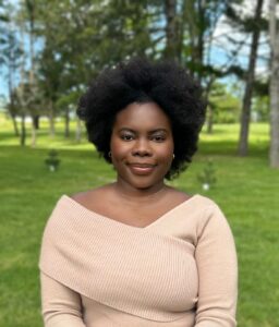 Smiling woman with dark brown skin, hair is an Afro, tan off the shoulder top? Park in background or greenery in background