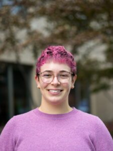 A headshot of Bella, who has short, curly hair dyed bright pink. She is wearing glasses with a clear frame and has a small, gold septum piercing. Bella is smiling and dressed in a light purple sweater. The background is slightly blurred, featuring some trees and a building.