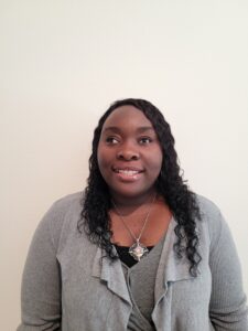 The picture shows Z'Leah standing against a plain, light-colored wall. She has dark skin and long, curly black hair. She is smiling and looking directly at the camera. She is wearing a gray cardigan over a black top and a large, ornate silver necklace with a central pinkish stone surrounded by smaller white stones. Her makeup is natural, enhancing her features subtly.