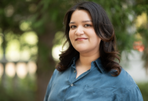 Alondra is a Latina woman with brown hair and brown eyes. She is wearing a blue satin button-up top.