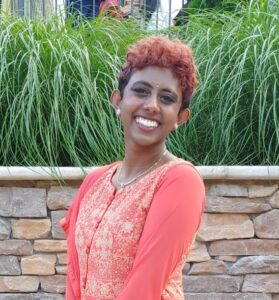 Dark skinned, Indian person with short orange hair, wearing a peachy orange kurta top. In front of brick and grass.