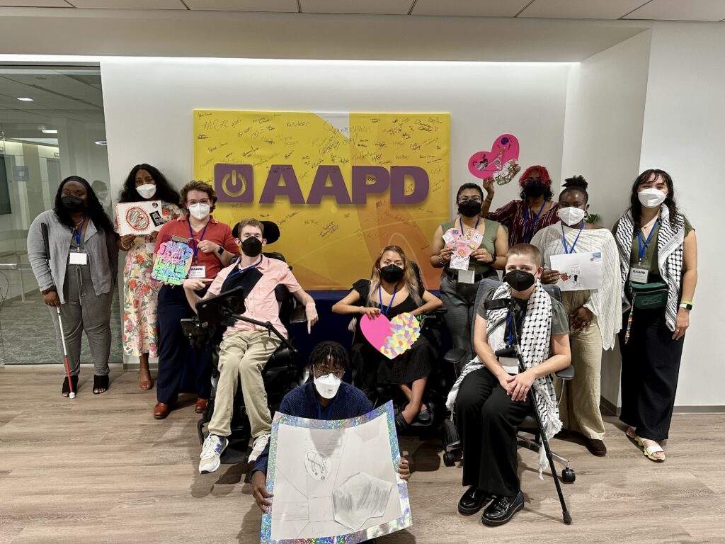 A group of diverse disabled interns - a young group of 10 disabled young people of a variety of races and disabilities, using a variety of mobility aids. The group is in the AAPD office, in front of a yellow sign with purple text that says "AAPD" and has its logo. The group is holding various crafts it made together.